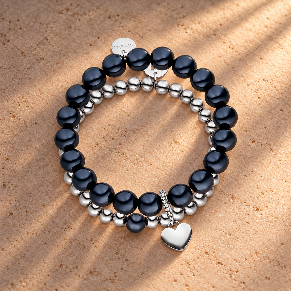 Perlenarmband Set mit Herzanhänger Schwarz - 3D Intensiv Glow Effekt & Versilbert – auf champagnerfarbenem Hintergrund im warmen Abendlicht.